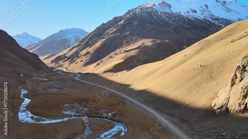 Aerial view of a mountain road running through a valley along a river, surrounded by mountains with snowy peaks. The road passes at an altitude of over 3500 meters above sea level and leads to the his