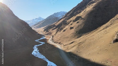 Aerial view of a mountain road running through a valley along a river, surrounded by mountains with snowy peaks. The road passes at an altitude of over 3500 meters above sea level and leads to the his