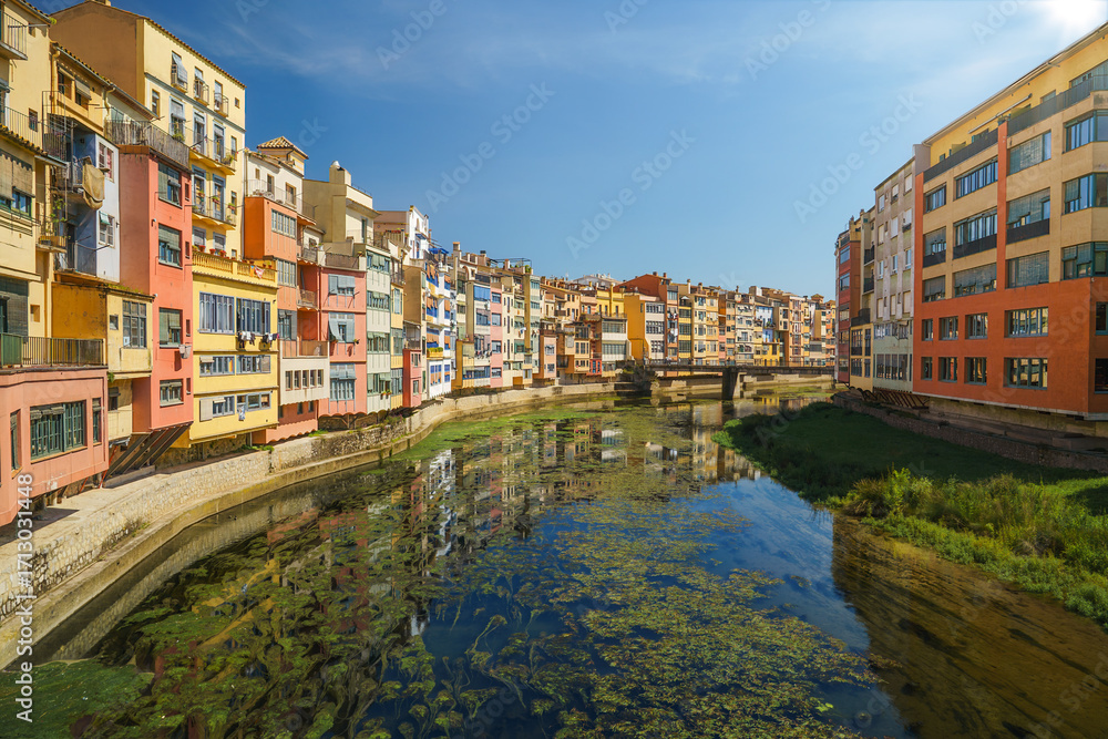 Fototapeta premium Colorful houses mirrored in the calm river Onyar, Girona’s Barri Vell under blue summer sky, Florentine feel; ideal for travel and city branding.