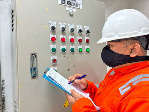 Industrial worker inspecting control panel with switches and indicators checking readings on clipboard ensuring safety protocols following procedures maintaining operational integrity