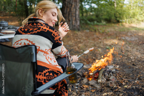 Camper woman resting in chair with drink near forest campfire