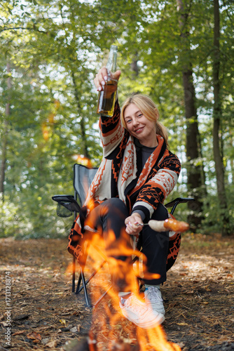 Woman enjoying camping with beer and roasting sausage by campfire in forest