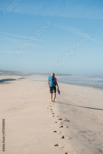 Fotografi Lonely barefoot backpacker man walking by sandy Portugese beach in morning on the Atlantic ocean coast