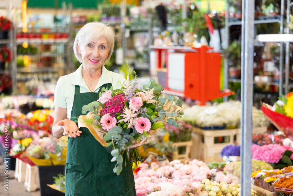 Fototapeta premium Positive experienced mature female florist checking and preparing fresh bouquet with roses and gerberas in floral shop