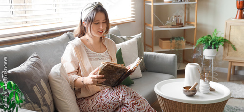 Fotografie Young witch with spell book sitting on sofa at home