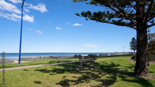 Park bench under tree shade overlooking Werribee South Beach in Victoria, Australia, with blue ocean, sandy shoreline, and grassy foreshore reserve. Concept of leisure, relaxation, coastal lifestyle