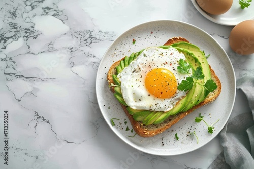 Top view of tasty breakfast toast with avocado and fried egg on a white plate with eggs on a marble background
