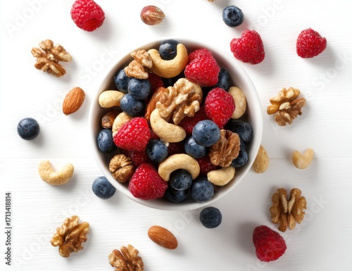 A white bowl brimming with blueberries, raspberries, walnuts, cashews, and almonds, scattered similarly around it on a white background, viewed from above