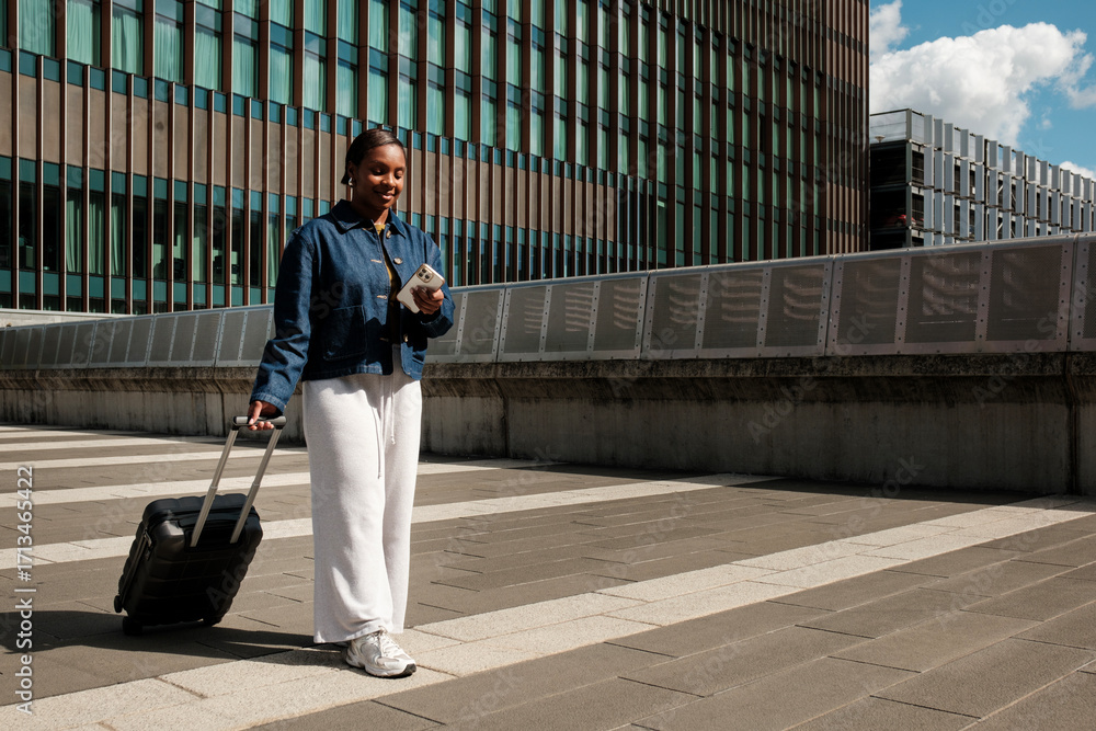 Fototapeta premium Young black woman using smartphone while pulling luggage in city