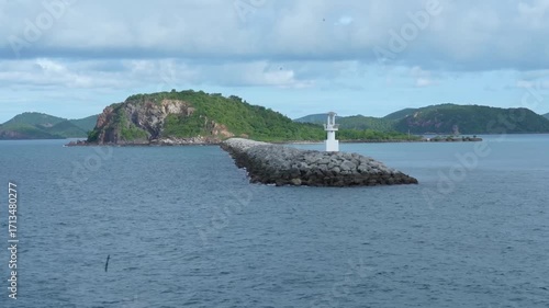 Rocky breakwater with small white lighthouse and calm blue sea, lush green island, distant hills, cloudy sky, peaceful coastal landscape, nature, rock, cloud, summer, outdoor scenery
