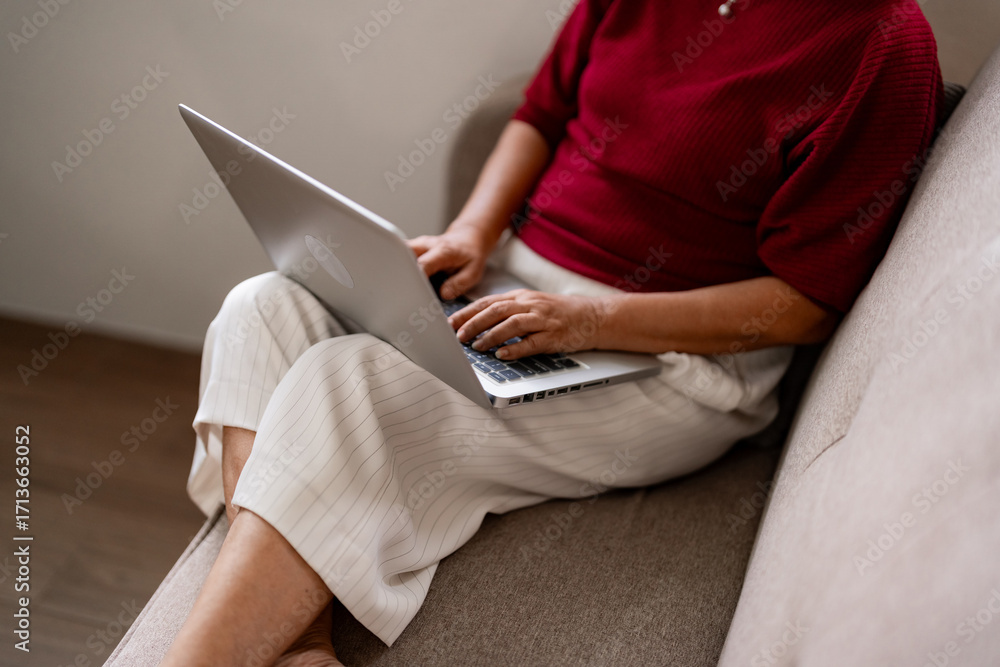 Naklejka premium Older Asian woman works from home using laptop computer on sofa modern living room digital connectivity