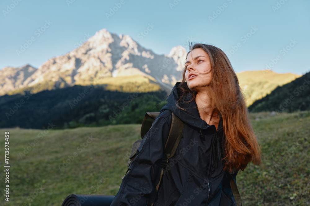 Fototapeta premium A solo hiker woman stands outdoors in a mountainous landscape. She carries a backpack and gazes toward the distant peaks with calm confidence and natural serenity.
