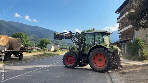 A green agricultural tractor uses its front loader to move large round hay bales on a sunny day. The scene is set in a picturesque rural area with rolling green mountains and clear blue skies.