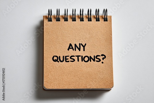 Tan notepad with black spiral binding and text ANY QUESTIONS on a white surface with shadows