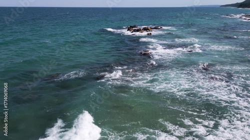 Wallpaper Mural Aerial view of waves crashing over coastal rocks on a turquoise sea, with a scenic sandy beach and forested cliffs in the background. Powerful water movement and beautiful seascape captured from above Torontodigital.ca