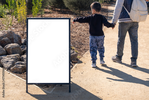 Blank white advertising mockup board on outdoor park pathway with an Asian child and adult parent walking hand in hand in the background. Concept of copy space, signage, family-friendly information.