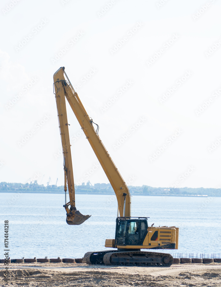 Φωτογραφία Excavator with bucket while working on the river bank against the background of blue sky and yellow sand pile