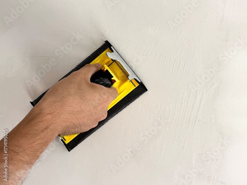 A builder smooths a wall after plastering using a sanding tool. The image depicts construction work and interior finishing in home improvement.