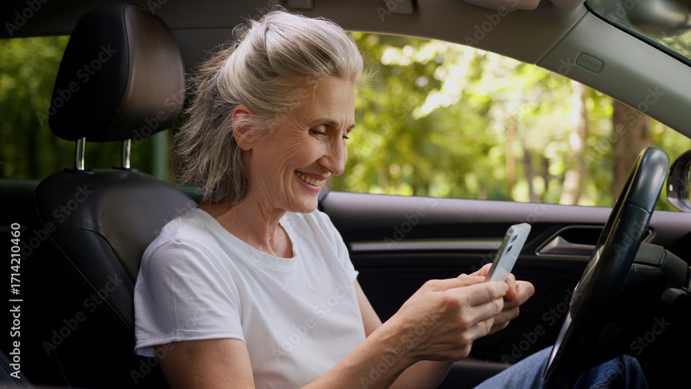 Naklejka premium Cheerful Caucasian mature woman elderly senior female retired pensioner car automobile driver inside auto using mobile phone playing game clench fist celebrating win victory betting success offer deal