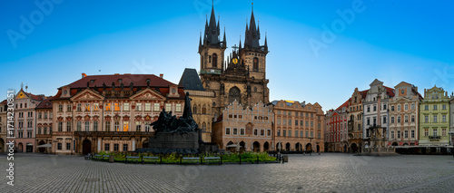 Panoramic view of Prague’s Old Town Square with Gothic Týn Church, Jan Hus Memorial, and historic architecture under a clear blue sky on an early summer morning day.