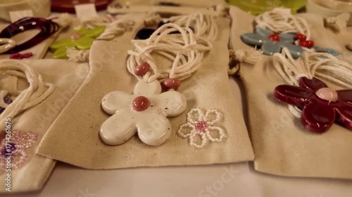 Close-up of handmade ceramic flower pendants on fabric pouches. Colorful artisanal jewelry with purple, white, and green designs, showcasing craftwork, creativity, and decorative accessories