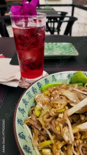Close-up of Pad Thai with chicken, lime, peanuts, and vegetables in a decorative bowl, served with a glass of red lemonade. Traditional Asian meal with vibrant colors and textures