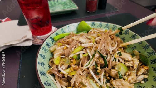 Close-up of Pad Thai with chicken, lime, peanuts, and vegetables in a decorative bowl, served with a glass of red lemonade. Traditional Asian meal with vibrant colors and textures