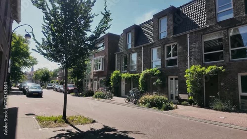 Sunny residential street with traditional Dutch brick houses, bicycles by the facades, and green plants decorating the entrances. Peaceful urban neighborhood