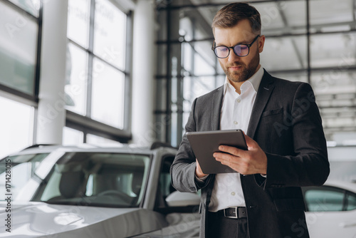 Fotografie Bearded friendly car seller in glasses and suit standing in car salon and holdin
