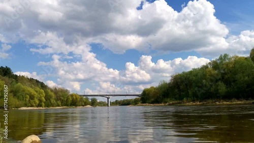 Timelapse of white clouds moving fast across blue sky over river and car bridge. Low view of flowing stream and traffic.