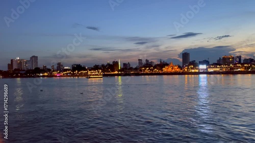 Scenic view of Phnom Penh riverside skyline at twilight, with illuminated buildings, temple architecture, boats, and colorful reflections shimmering on the Tonle Sap River