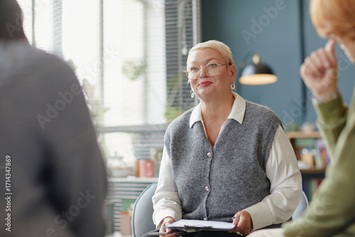 Caucasian woman wearing glasses leading group discussion with two people in modern office setting, holding clipboard and smiling while engaging participants
