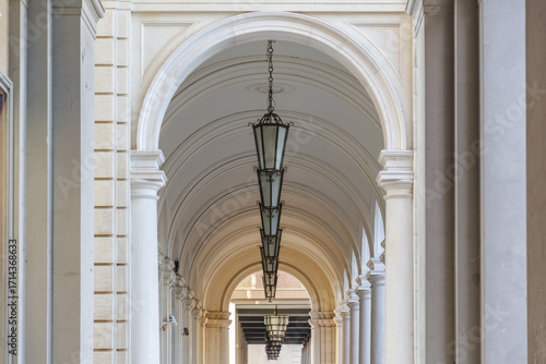 View of a long corridor, defined by light-colored arches and columns, with hanging lanterns casting soft shadows in a symmetrical display, Chieti, Abruzzo, Italy.