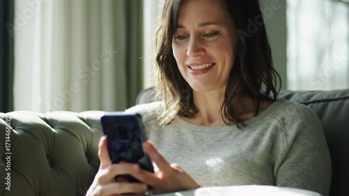 Happy casually dressed woman sitting on cozy sofa with modern smartphone in hands. Attractive caucasian brunette smiling while looking on mobile scree