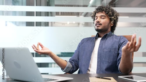 Young businessman meditating with closed eyes during a break while sitting at workplace in business office. Calm handsome worker practices yoga and feels peace of mind. Male relaxes to relieve stress