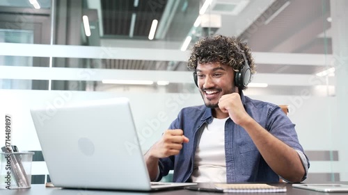 Excited joyful young businessman in headphones watching sports match or competition using laptop sitting at workplace in business office. Happy man cheering for bids at auction, celebrating success