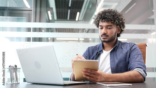 Young businessman watching video call conference or training takes notes looking at laptop screen sitting at workplace in business office. Focused worker listening to remote online course, seminar