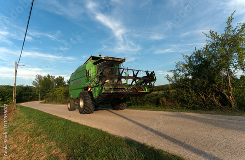 A vibrant green combine harvester, seen from a low angle, drives along a winding rural road under a bright blue sky with wispy clouds, flanked by lush green foliage.