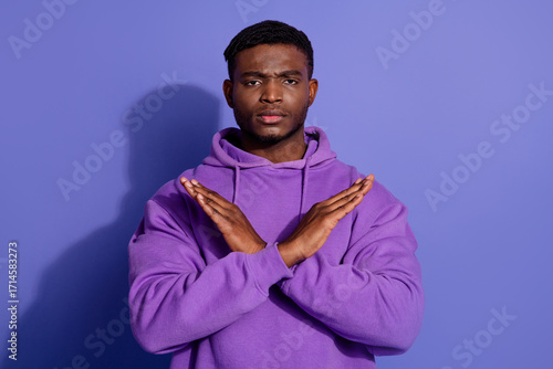 Confident young man in casual sportswear crossing arms to signify refusal against purple background