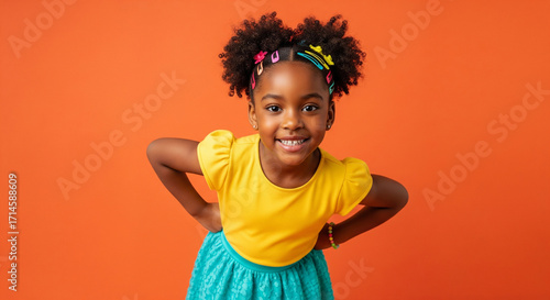 A cheerful and happy Black girl wearing bright yellow and blue clothes with vibrant hair clips, posing confidently against a solid orange studio background