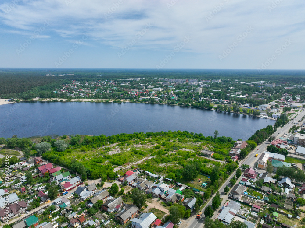 Obraz premium A wide aerial view of Gus Khrustalny, showing residential houses along the shore of a large lake with green fields and a dense forest on the horizon.
