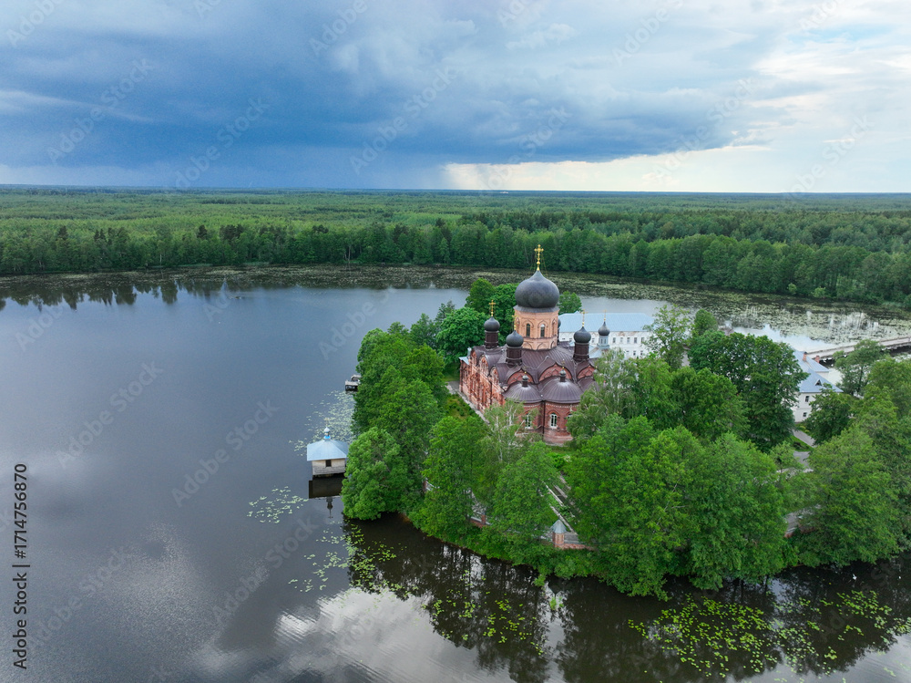 Obraz premium An aerial view of the Vvedensky Island Monastery with its black domes and red walls, located on a small, tree-covered island in a wide lake.