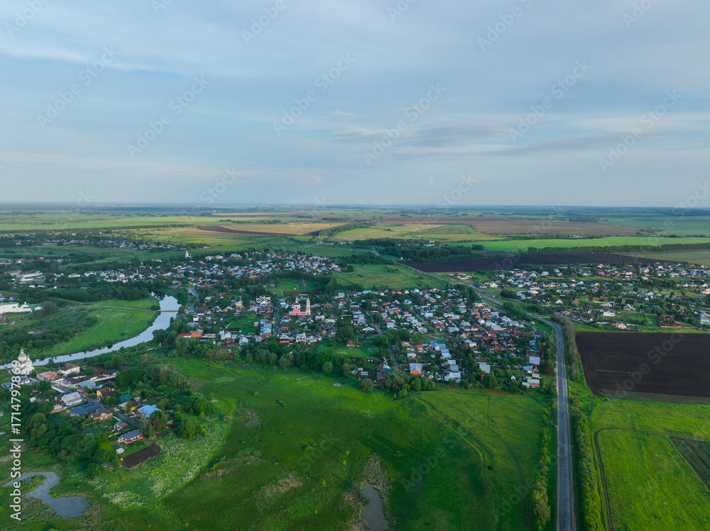 Fototapeta premium A wide aerial shot of the city of Suzdal, showing a mix of residential houses and a road, surrounded by vast green fields and forests.