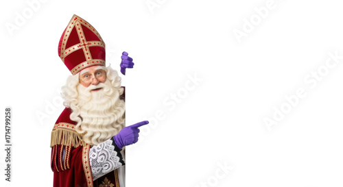 Elderly caucasian Sinterklaas with long white beard, opulent red and gold robe, purple gloves, peeking from large white sign, pointing to copy space, gentle studio portrait concept for holiday