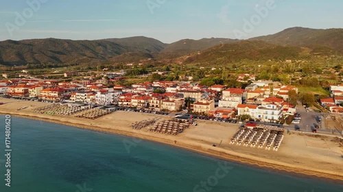 High-resolution aerial drone image showcasing a picturesque beachfront resort scene. Neatly arranged white umbrellas and lounge chairs line the golden sandy shore, while colorful hotel buildings with 