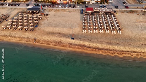 High-resolution aerial drone image showcasing a picturesque beachfront resort scene. Neatly arranged white umbrellas and lounge chairs line the golden sandy shore, while colorful hotel buildings with 