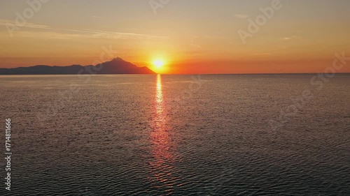 Breathtaking aerial drone capture of a tranquil sunset over a peaceful sea, with the sun dipping behind a distant mountain range. The sky glows in rich shades of orange and yellow, transitioning into 