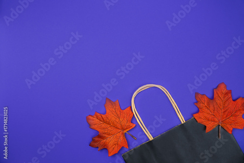 Black paper shopping bag and autumn leaves on an purple background. Autumn shopping and sales concept. Top view. Flatlay. Copy space.