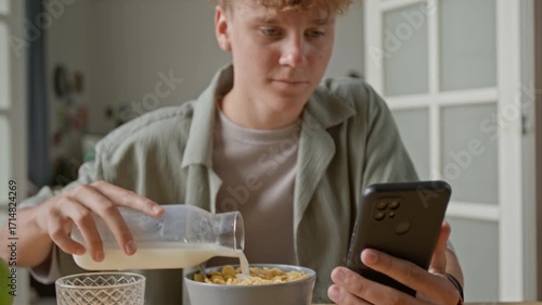 Tilt up shot of young man with curly hair watching videos during breakfast focus on hand holding smartphone, copy space
