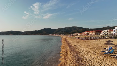 High-resolution aerial drone image showcasing a picturesque beachfront resort scene. Neatly arranged white umbrellas and lounge chairs line the golden sandy shore, while colorful hotel buildings with 